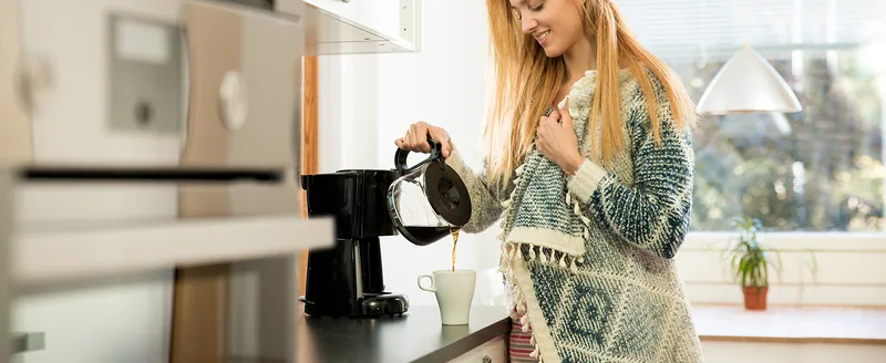 A woman in a knit cardigan pours coffee from a drip coffee maker carafe in a bright kitchen