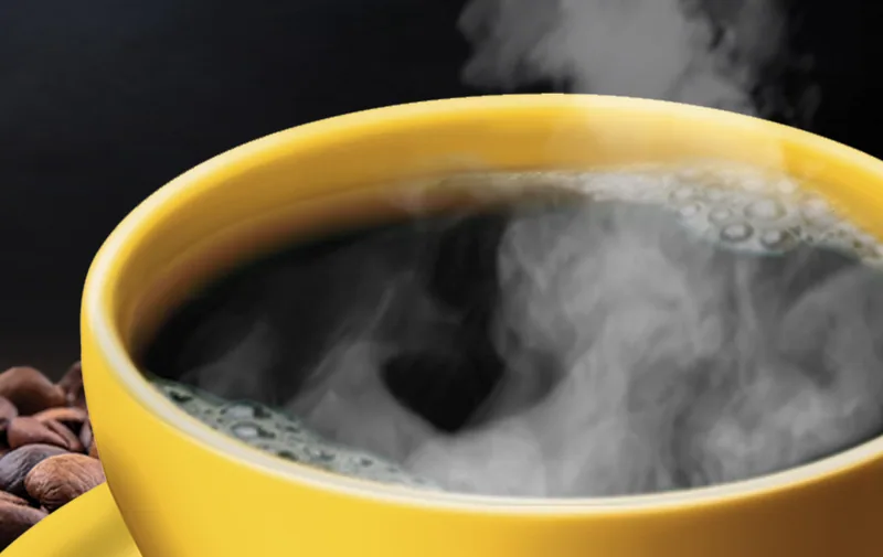 Steam rises from a yellow cup of black coffee beside scattered roasted beans
